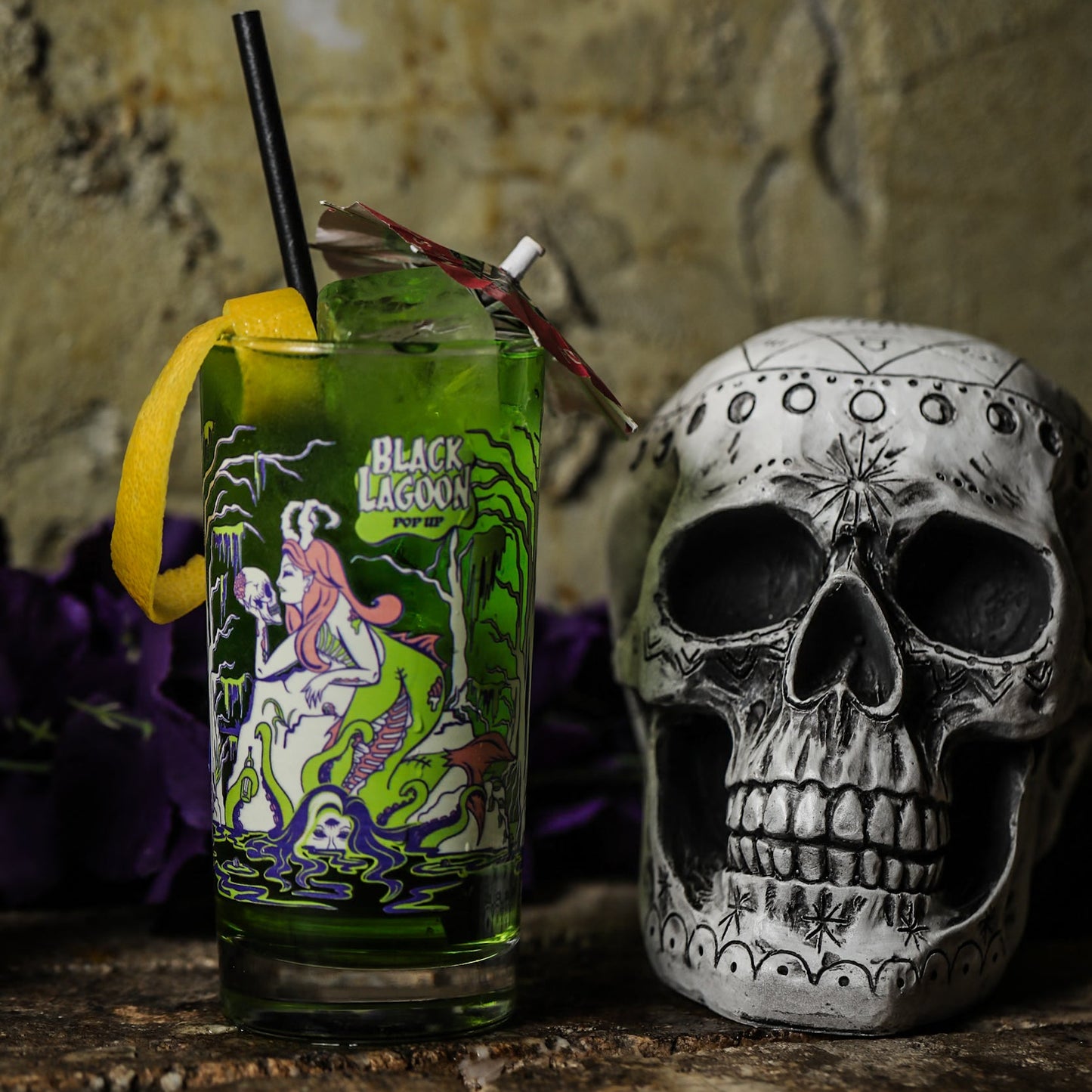 Black Lagoon cocktail glass with a zombie mermaid printed on next to a skull on a rustic surface.