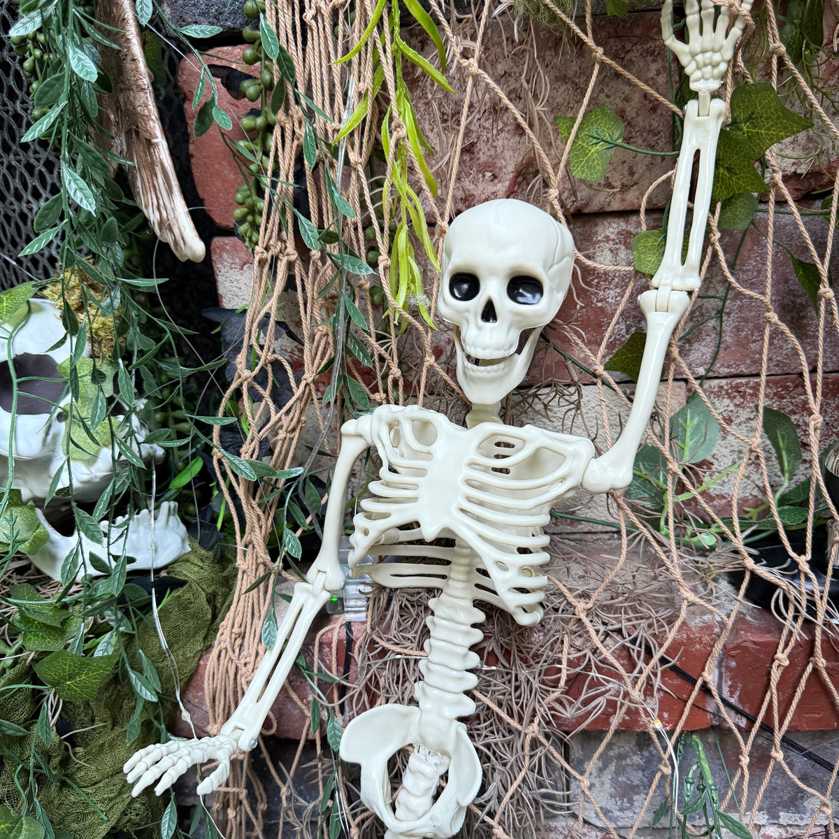 An articulated plastic skeleton decoration posed against a rustic brick wall backdrop with hanging green foliage and netting.